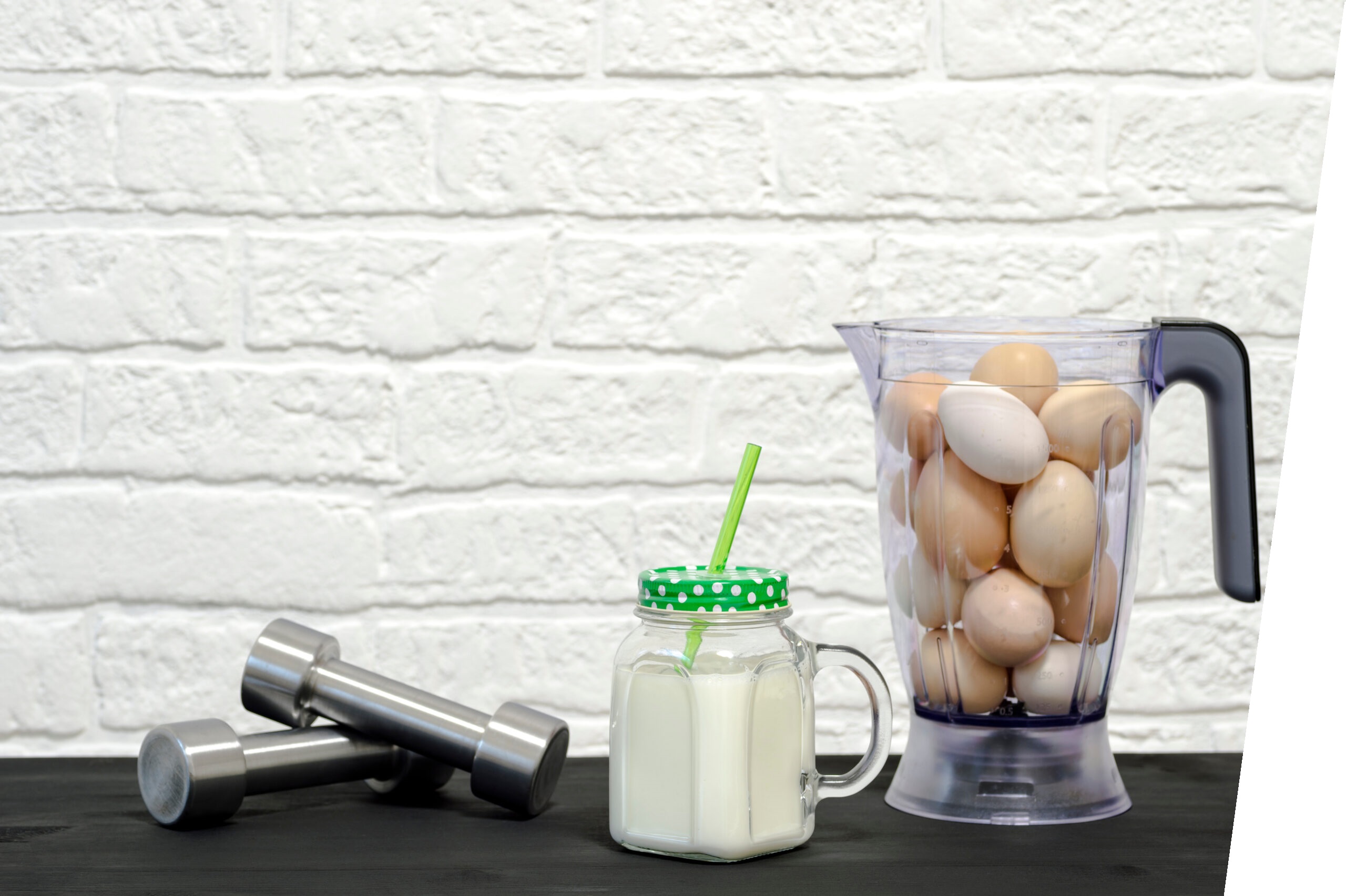 Protein shake in a mug against the background of a blender bowl with chicken eggs and dumbbells against a white wall.